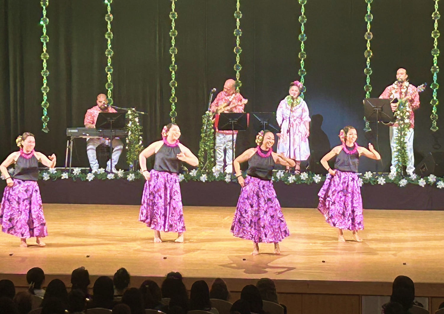 ステージで踊る4名の女性 紫色のスカートに黒色のノースリーブ バックには演奏するバンド